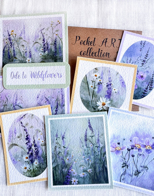 A collection of handmade floral art cards featuring watercolor wildflower designs in shades of purple, blue, and white, arranged on a textured white fabric background. One card reads 'Ode to Wildflowers' and another brown card reads 'Pocket ART collection'.