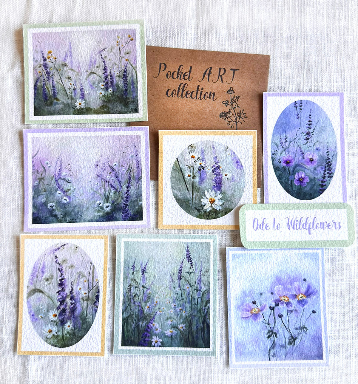 A collection of watercolor floral pocket art cards featuring lavender, purple, and white wildflowers, arranged on a light gray textured fabric background. A brown card in the center reads 'Pocket ART collection', and another card says 'Ode to Wildflowers'.
